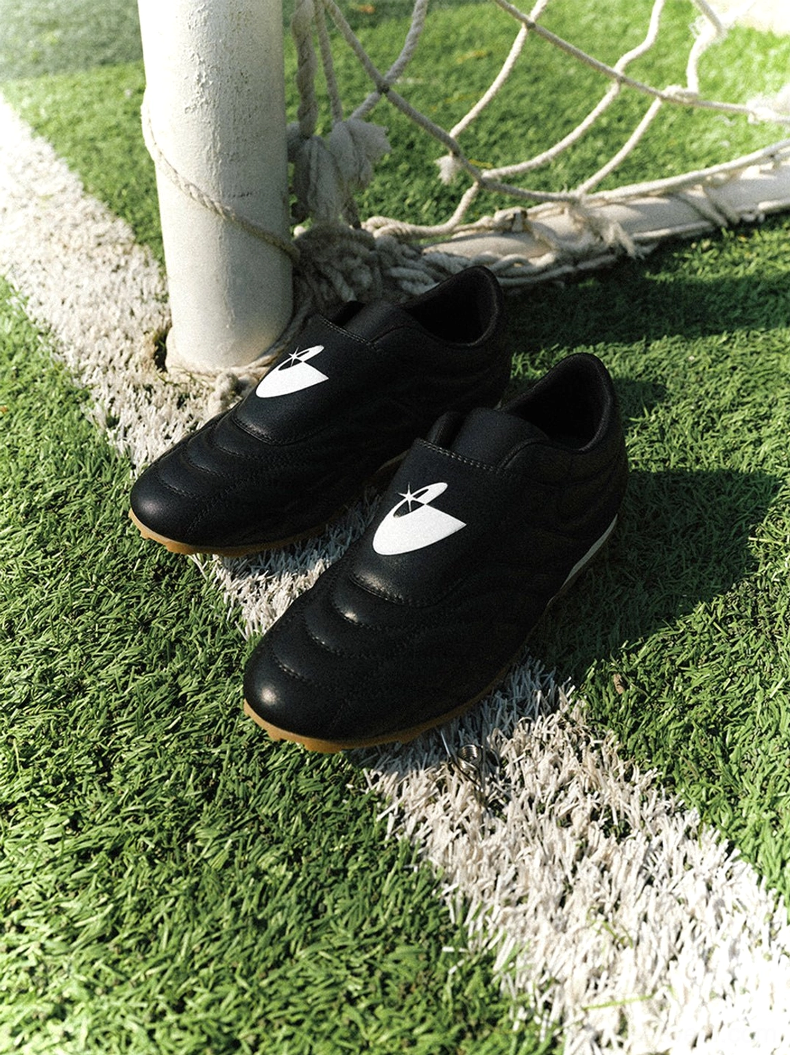 Pair of black football-inspired trainer sneakers placed beside a goal post on artificial turf in natural sunlight