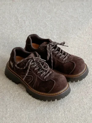 Pair of dark brown chunky suede sneakers displayed from an angled top-down view on a light grey carpet with matching laces, contrast stitching, and ridged rubber outsoles