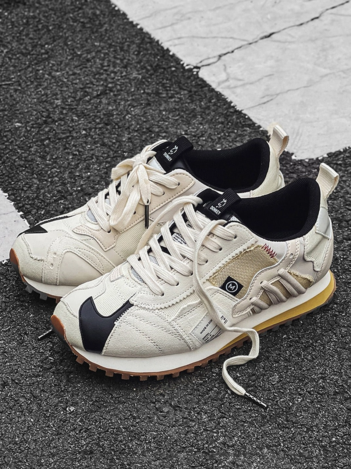 Pair of ivory retro runner sneakers with black toe caps, lace-up design, layered panels, and brown gum waffle soles on asphalt