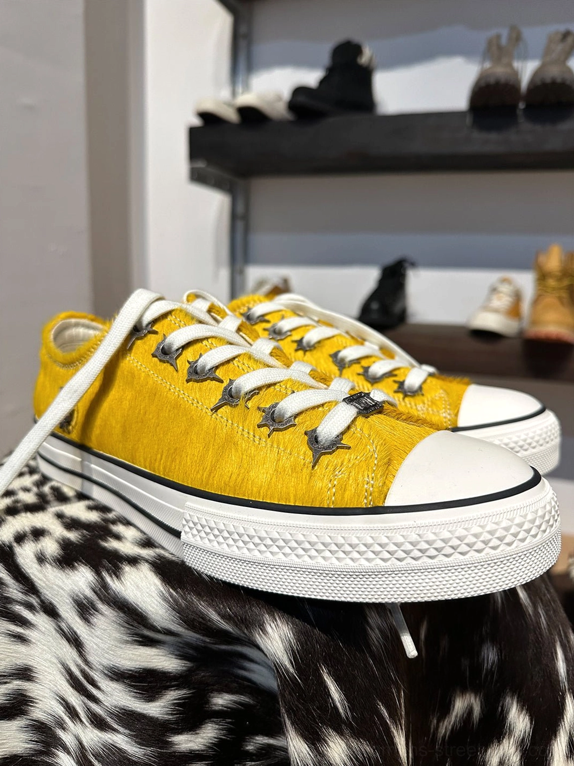 Pair of mustard yellow cowhide low-profile sneakers displayed on a spotted cowhide background with contrast laces and rubber toe caps