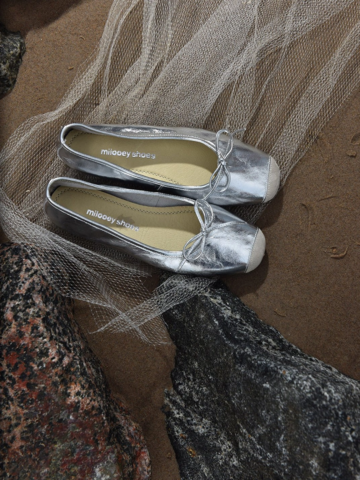 Pair of silver metallic square-toe bow ballet flats displayed on sand with natural rocks