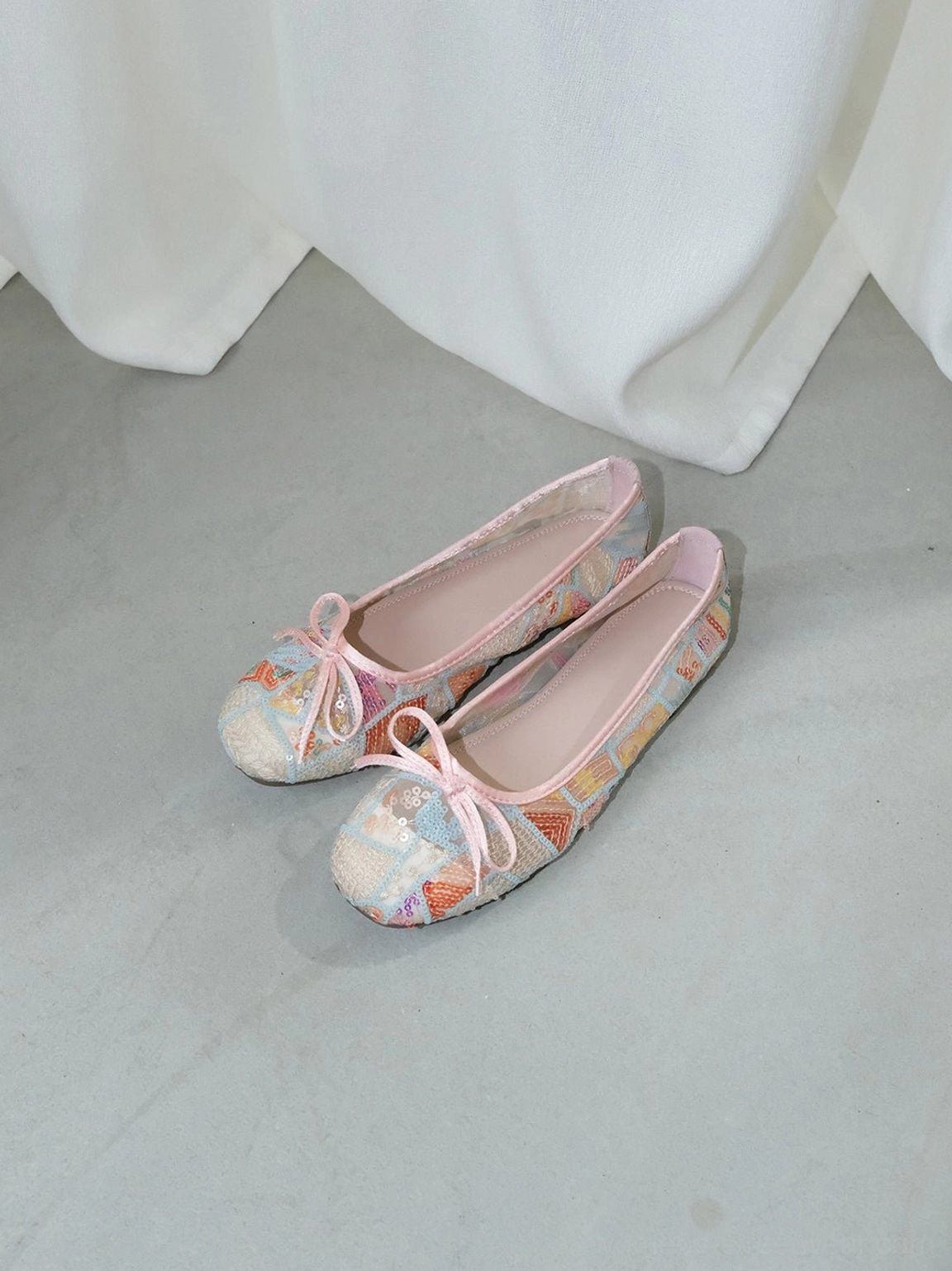 Pair of pastel multicolor floral mesh bow ballet flats with pink trim displayed on a light gray floor by a white curtain