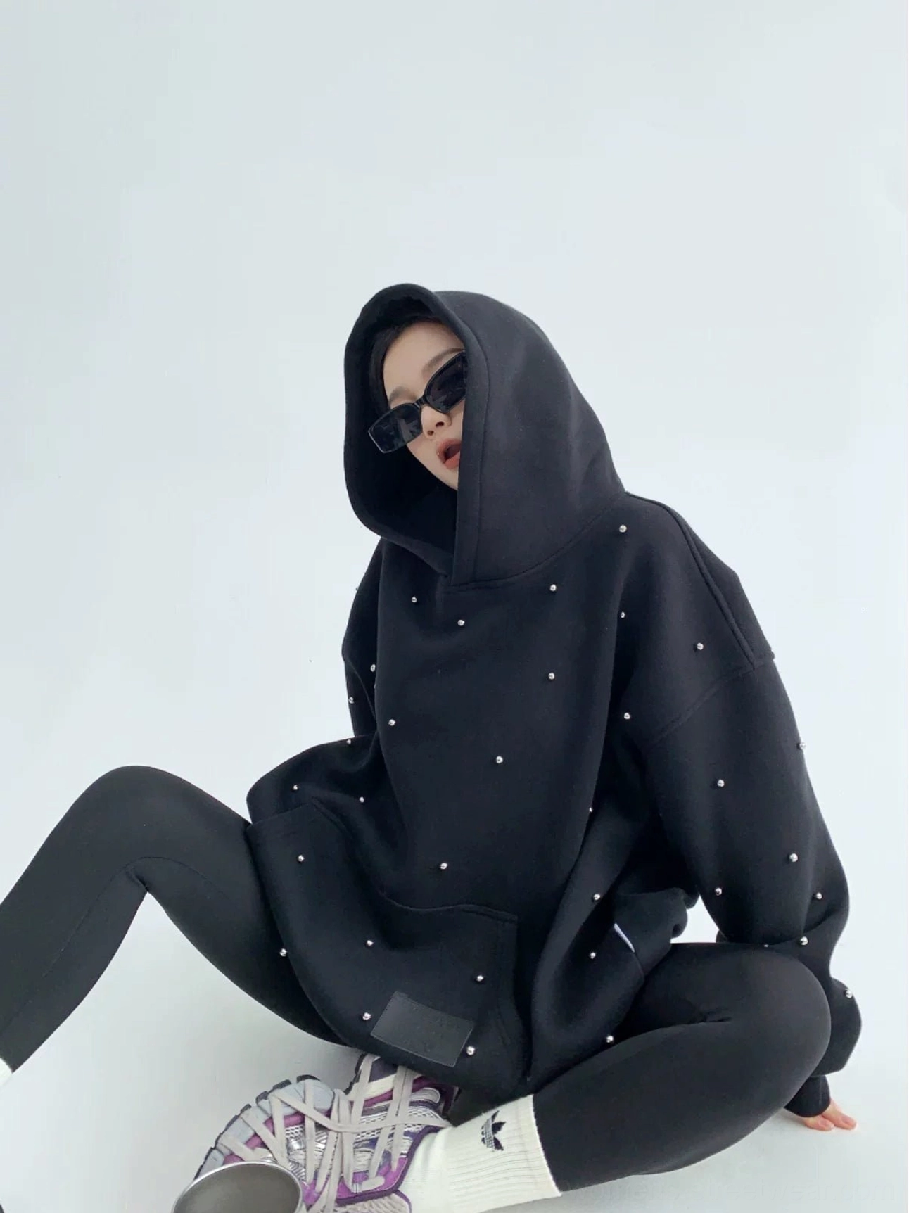 Black pearl-studded oversized streetwear hoodie on a seated female model with sunglasses, fitted leggings, sneakers, and a front kangaroo pocket