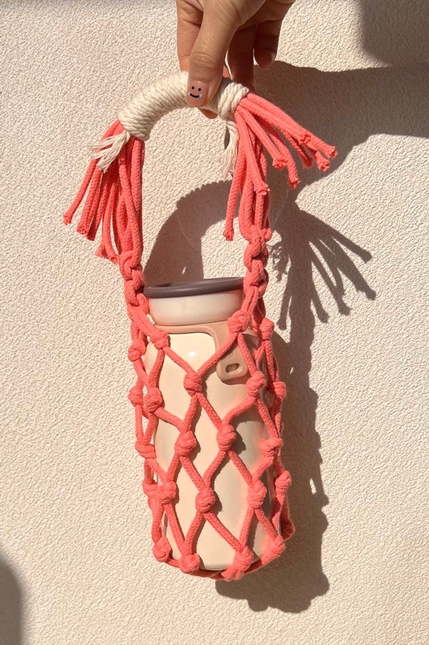Pink cotton macrame water bottle carrier holding a standard tumbler, shown with a decorative tassel handle and woven net pocket