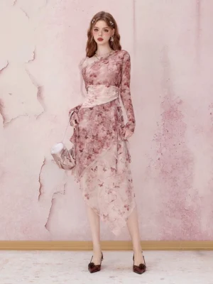 Full body front view of a woman wearing a pink floral asymmetrical mesh midi dress with long sleeves, a light pink waist sash, high heels, and a handbag