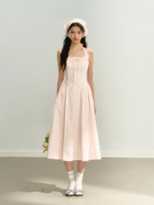 Front full-body view of a pastel pink halter bow midi dress with ruched bodice, gathered waist, flowing A-line skirt, matching headscarf, white socks, and silver flats
