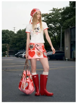 Full-body urban streetwear outfit featuring a pink kitty heart patchwork mini skirt, white cropped ringer tee, red rain boots, printed tote bag, and headscarf