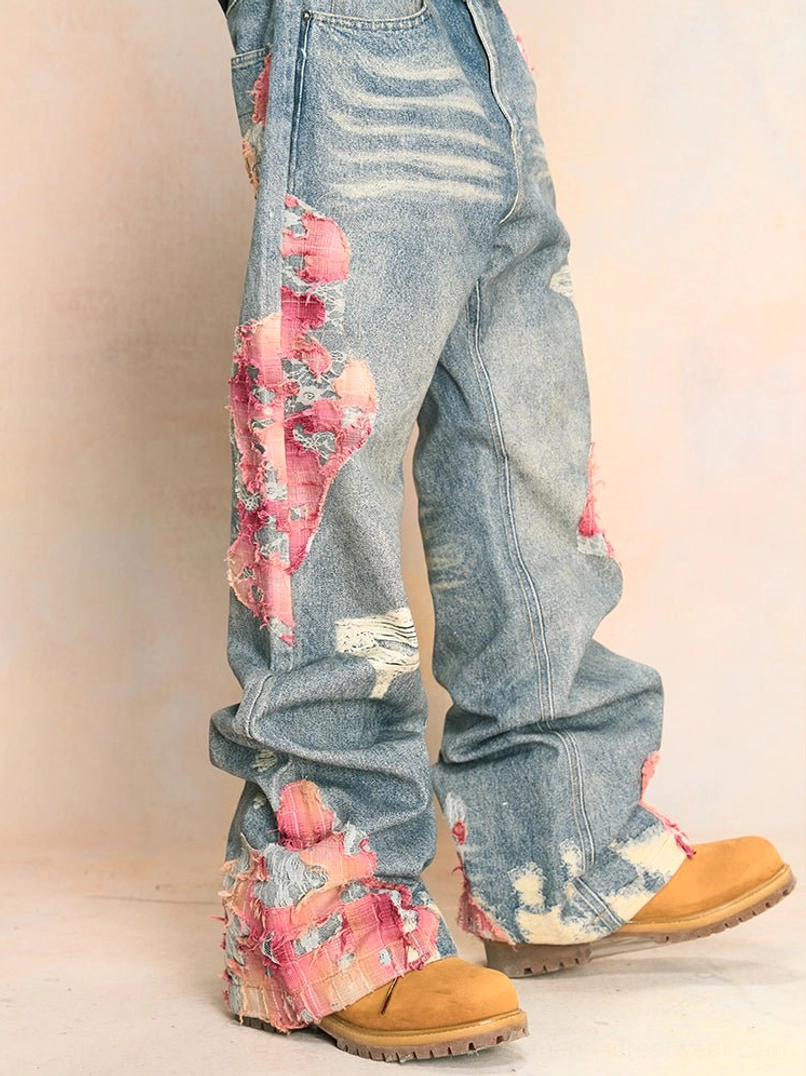 Low angle front three-quarter view of faded blue distressed wide-leg jeans with pink lace-style embroidery, frayed patches, ripped denim details, and tan work boots