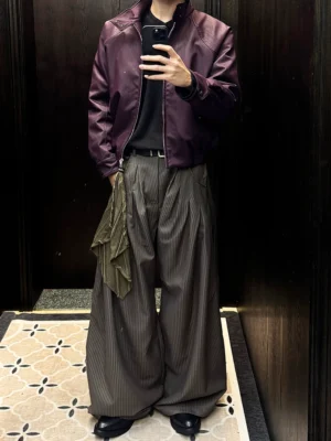 Model wearing pinstripe barrel leg streetwear pants with front pleats, draped tassel detail, cropped purple leather jacket, black belt, and leather shoes near a hotel elevator