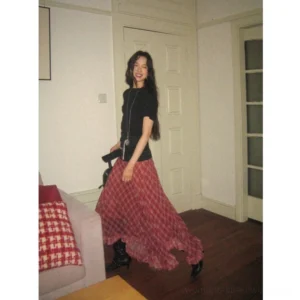 Red plaid chiffon asymmetric hem maxi skirt styled with a black short sleeve top and ankle boots in a vintage apartment