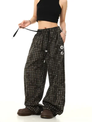 Left angle view of relaxed plaid wide-leg streetwear pants showing the adjustable drawstring waist decorative leg badges and roomy silhouette