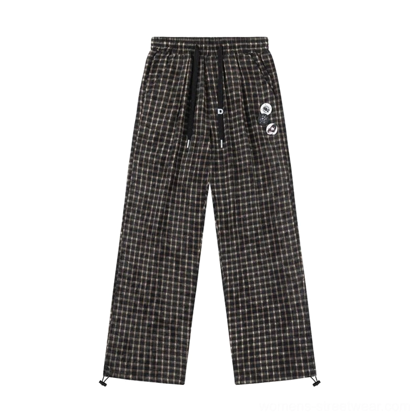 Flat lay view of plaid wide-leg streetwear track pants on a white background showing the complete garment shape drawstring waist side pockets and decorative badges