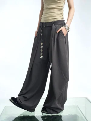 Dark gray pleated balloon wide-leg streetwear pants shown from a three-quarter side view with flowing draped panels, high waist, detachable geometric chain, cropped beige top, and platform shoes