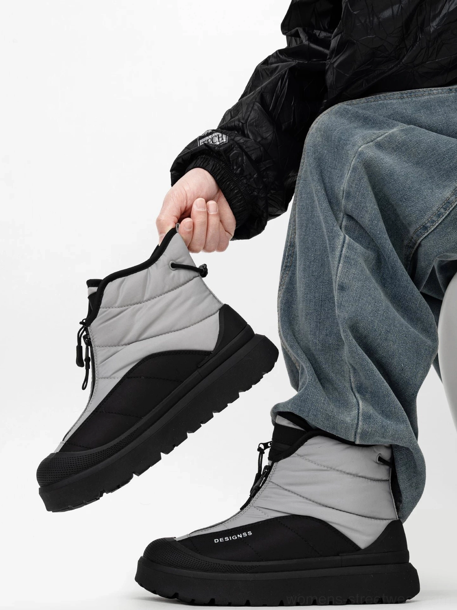 Three-quarter view of black and gray zip-up snow boot held by model, featuring padded ankle collar, front zipper closure, and matte platform outsole