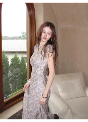Porcelain blue floral column maxi dress shown from a left three-quarter view on a model in a vintage-inspired living room with a white sofa and greenery