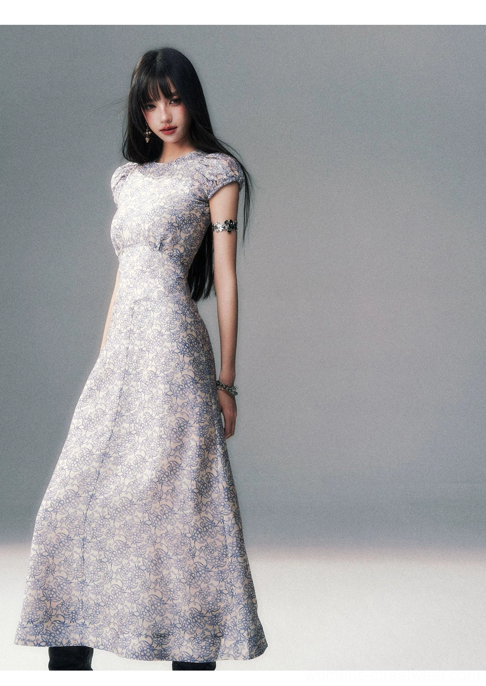 Porcelain blue floral fitted maxi dress shown from the left three-quarter view with a defined waist, flowing long skirt, and silver arm jewelry