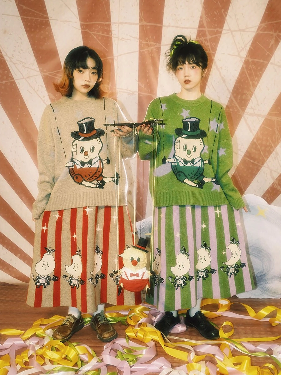 Two models wearing colorful puppet swing piglet jacquard knit sweaters with coordinated printed skirts against a retro circus backdrop