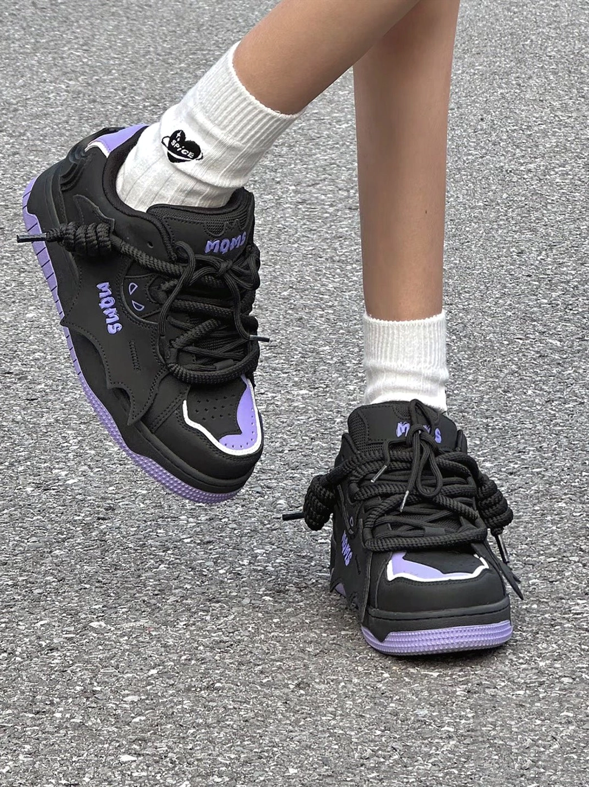 Black and purple chunky platform skate sneakers worn with white crew socks in an on-foot streetwear scene, showing the graphic side profile and exaggerated thick laces