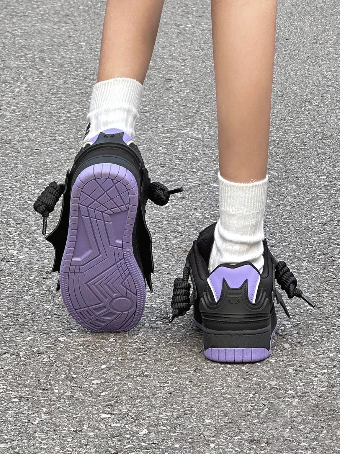 Rear and outsole view of black and purple bat graphic platform sneakers worn on pavement, featuring purple chevron rubber soles, wing-shaped heel details, and chunky skate styling