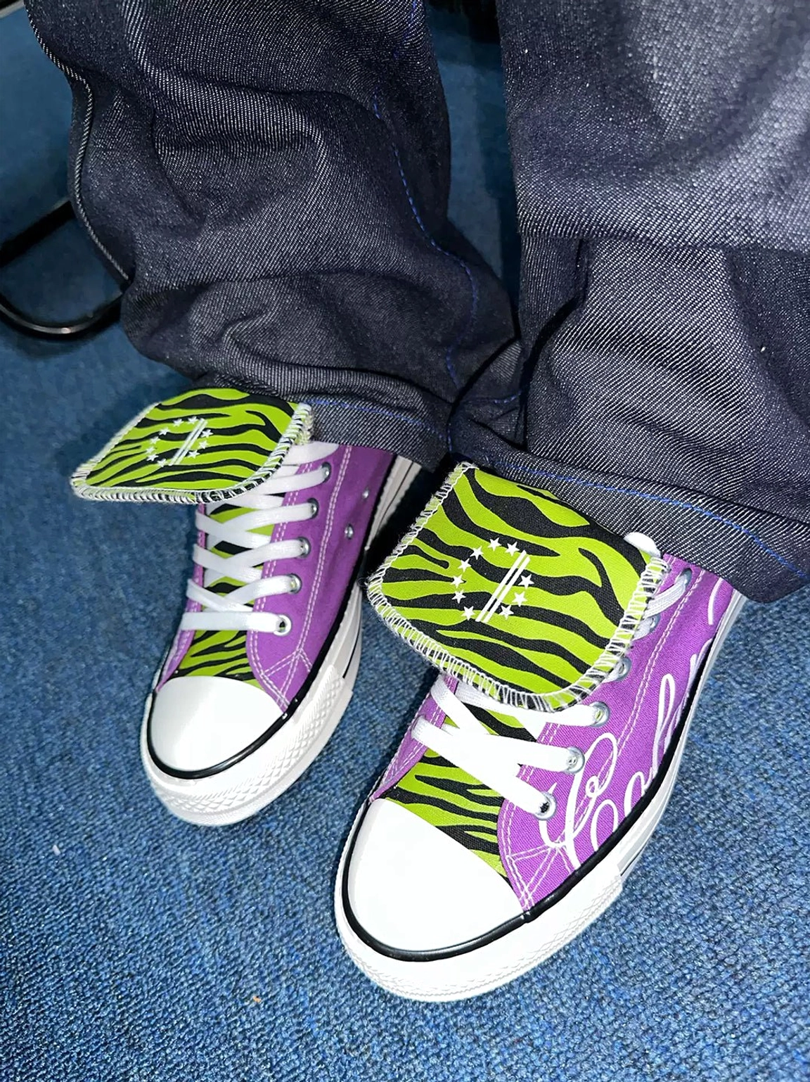 Angled view of purple punk canvas sneakers styled with dark denim, contrasting green zebra print tongues, white laces, and graphic side lettering