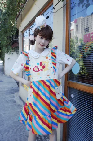 Three quarter side view of colorful rainbow stripe bear overall dress showing kawaii cartoon print, fitted bib silhouette, ruffle hem and matching hair bow