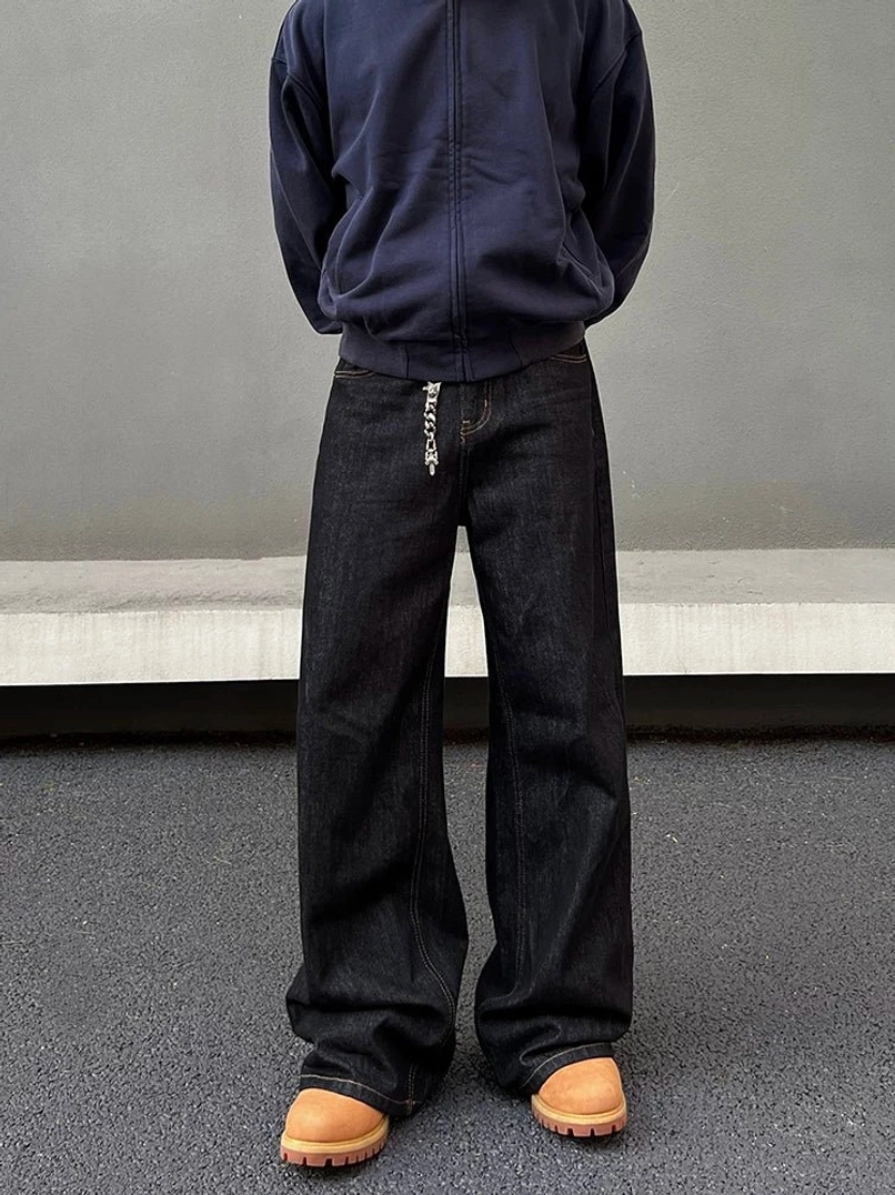 Front full-body view of dark indigo raw denim wide-leg jeans styled with a navy zip-up sweatshirt and tan chunky work boots in a minimalist streetwear setting
