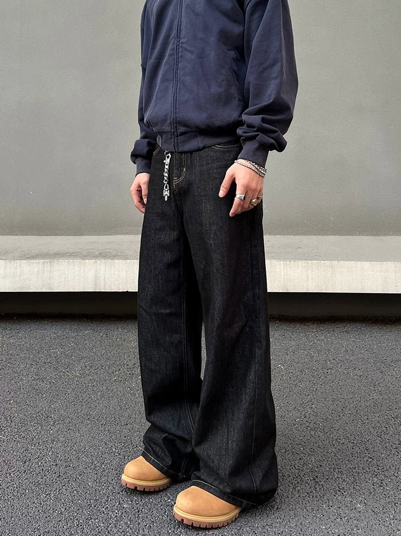 Three-quarter side view of wide-leg raw denim jeans showing the relaxed hip and thigh fit, deep indigo wash, waist chain, and chunky streetwear boots