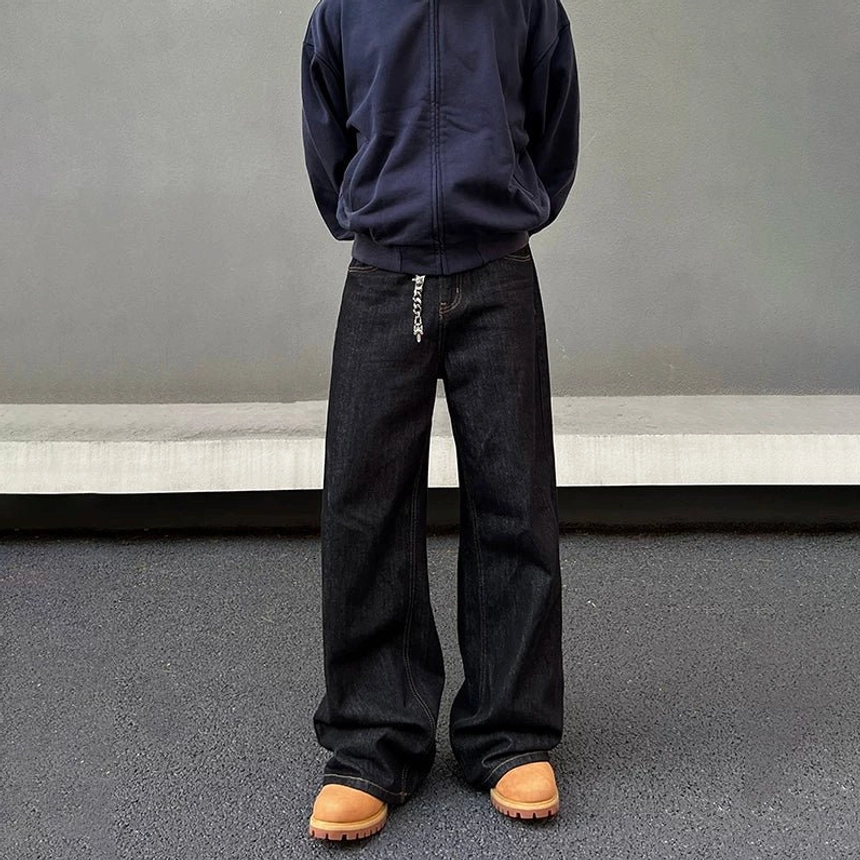 Close front-adjacent view of raw denim wide-leg jeans highlighting the deep indigo texture, loose silhouette, metal waist chain, and modern streetwear styling