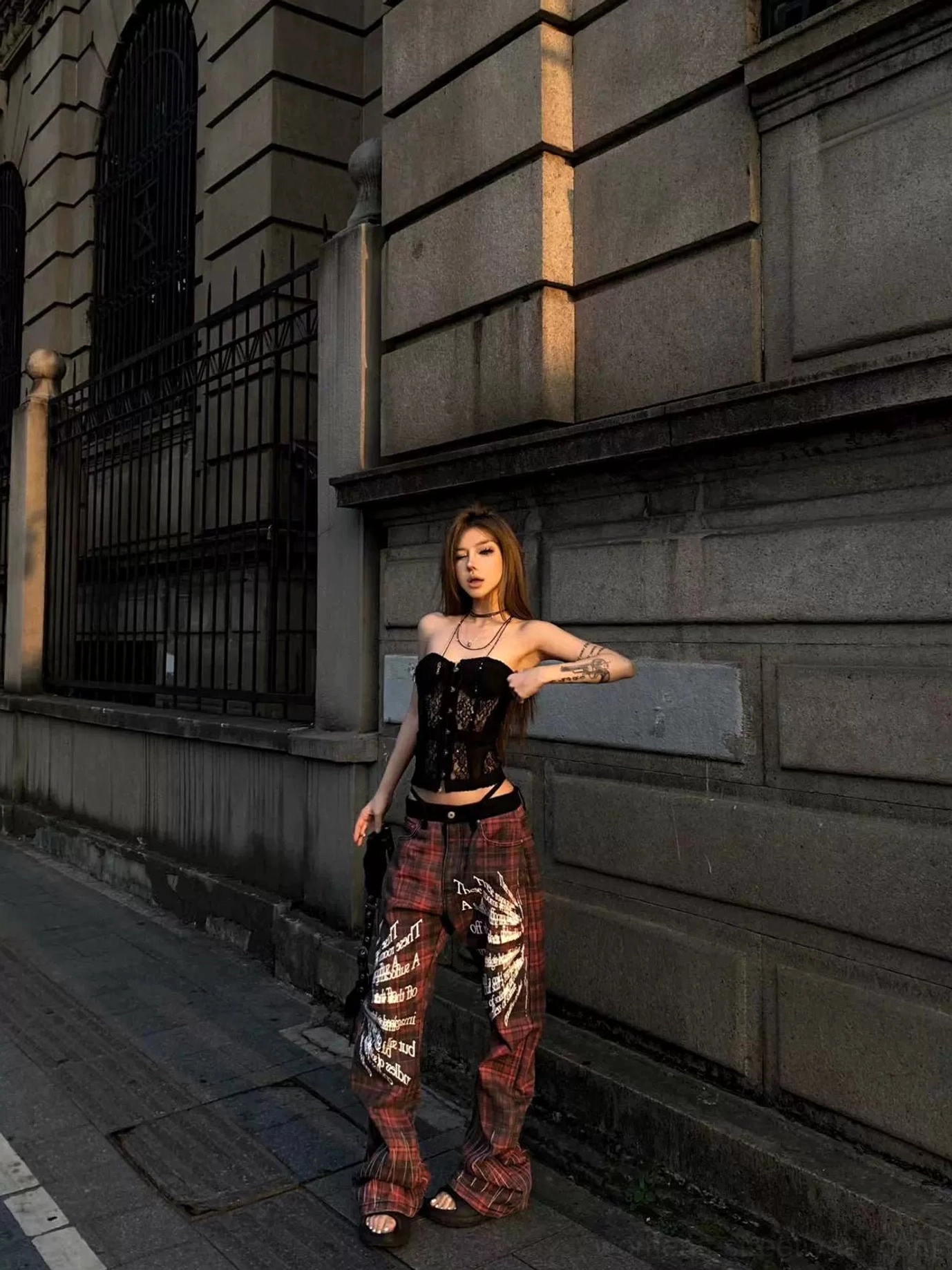 Front three quarter view of red black plaid gothic flare streetwear pants with white sigil lettering and wing graphics on a female model