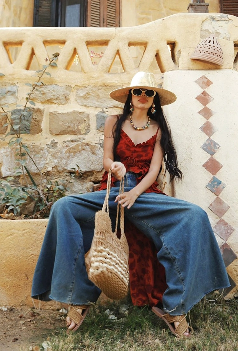 Red boho ruffle high-low camisole top styled with wide-leg jeans, straw hat, sunglasses, woven tote, and statement necklace outside a stone villa