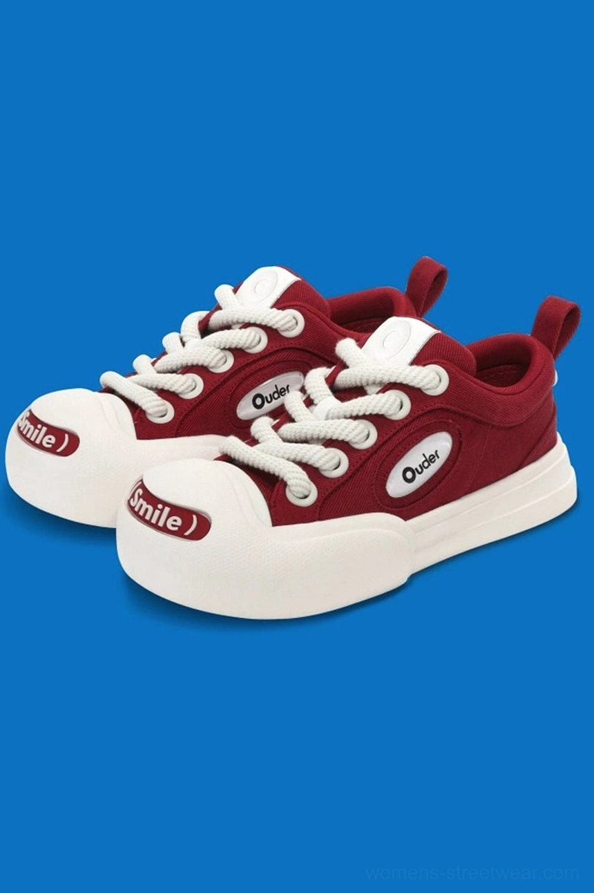 Side front view of red canvas low-top sneakers with off-white rubber sole and toe cap, Smile print, white laces, and oval side logo detail