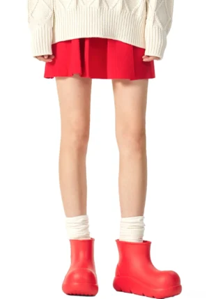 Model wearing red chunky ankle rain boots with a red mini skirt, cream knit sweater, and slouch socks for a Korean streetwear inspired outfit