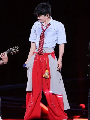 Red and grey detachable layered balloon parachute streetwear pants worn with a white shirt and red tie on a concert stage, showing oversized wide legs, drawcord details, pendant accents, and reflective lower-leg trim