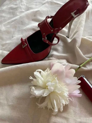 Red pointed-toe stiletto pumps with layered silver buckle straps displayed on off-white linen with peony flowers and lipsticks