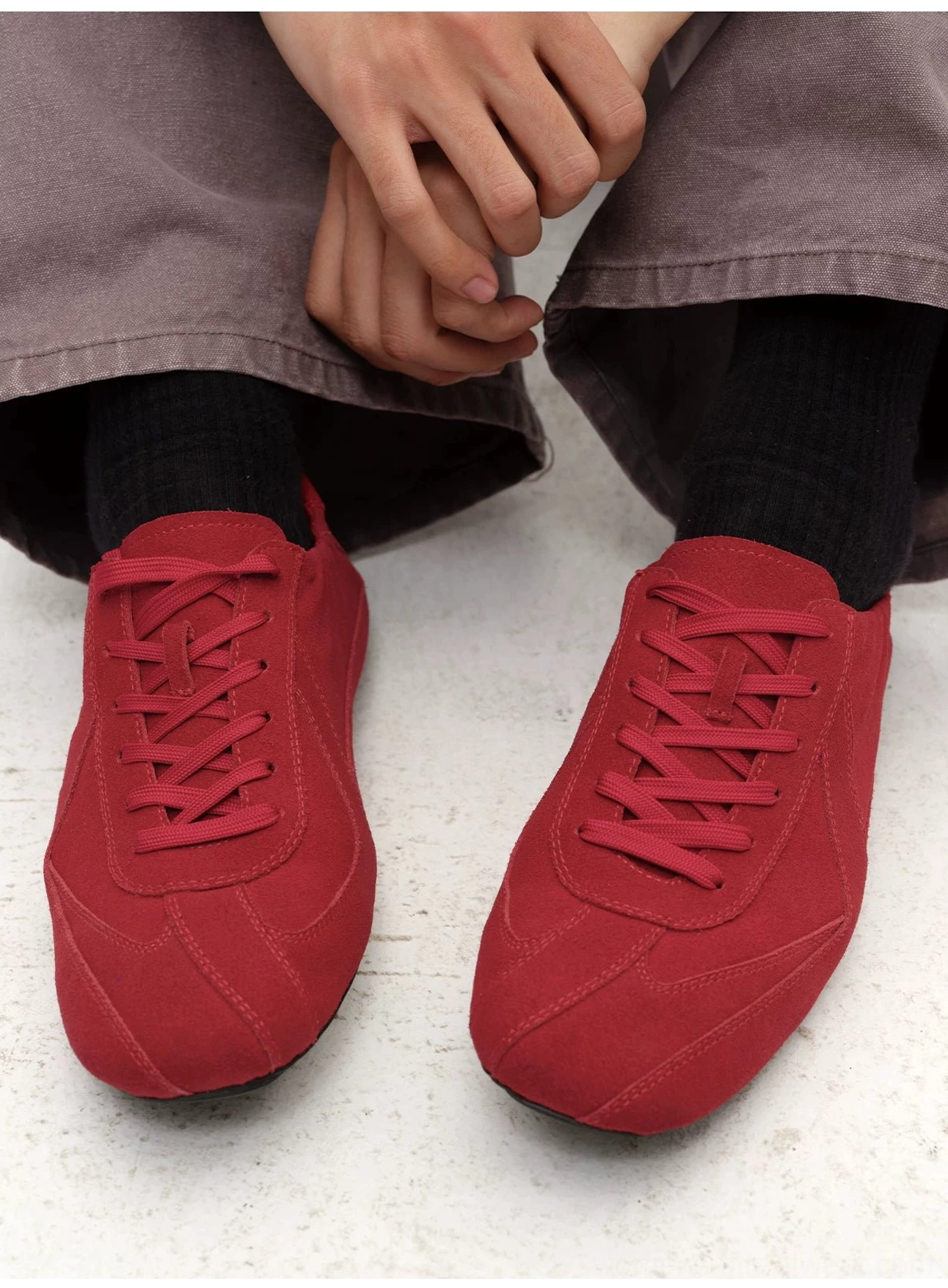Red suede minimalist lace-up sneakers worn with grey trousers and socks, showing a streamlined low-top streetwear fit