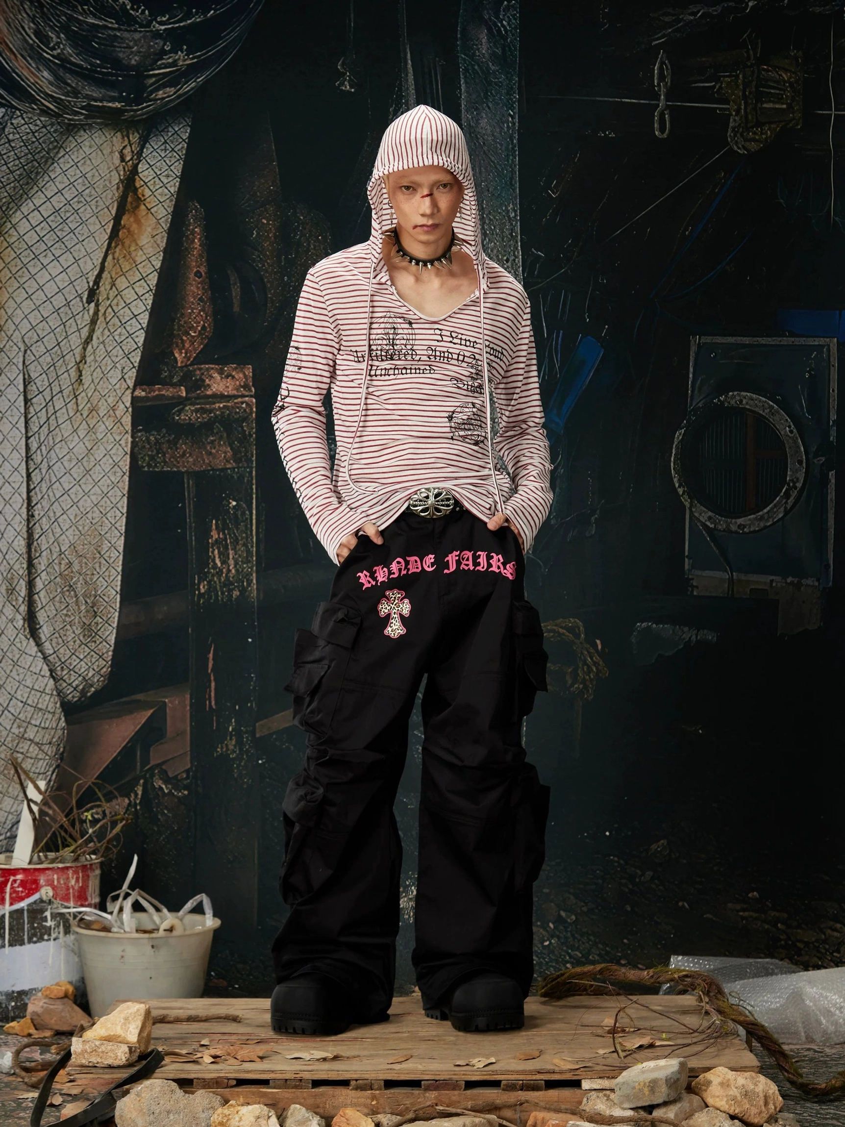 Model wearing a red and white striped gothic hooded long sleeve top with the hood up and black cargo pants with graphic lettering