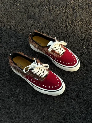 Reddish brown studded suede low-top sneakers shown from a top-down view on dark asphalt, featuring a wine red suede toe cap, fuzzy brown side panels, silver studs, white laces, and an off-white rubber sole