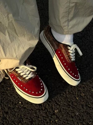 Reddish brown studded suede streetwear sneakers worn with beige loose cargo pants and white crew socks, showing the metallic toe embellishments and classic white lace-up design