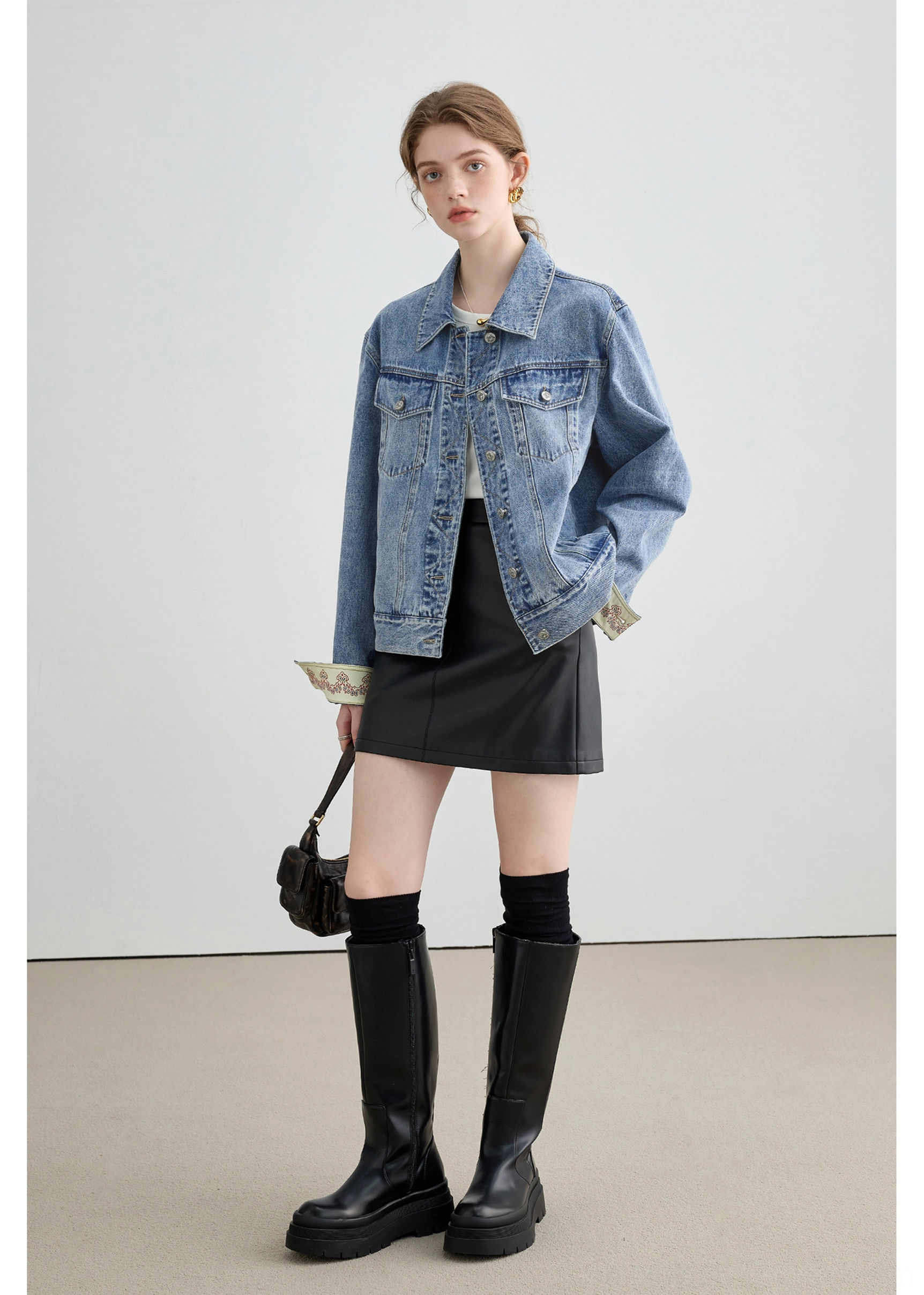 Full body side view of a female model wearing a relaxed light wash denim jacket with black mini skirt, knee-high boots, and mini bag