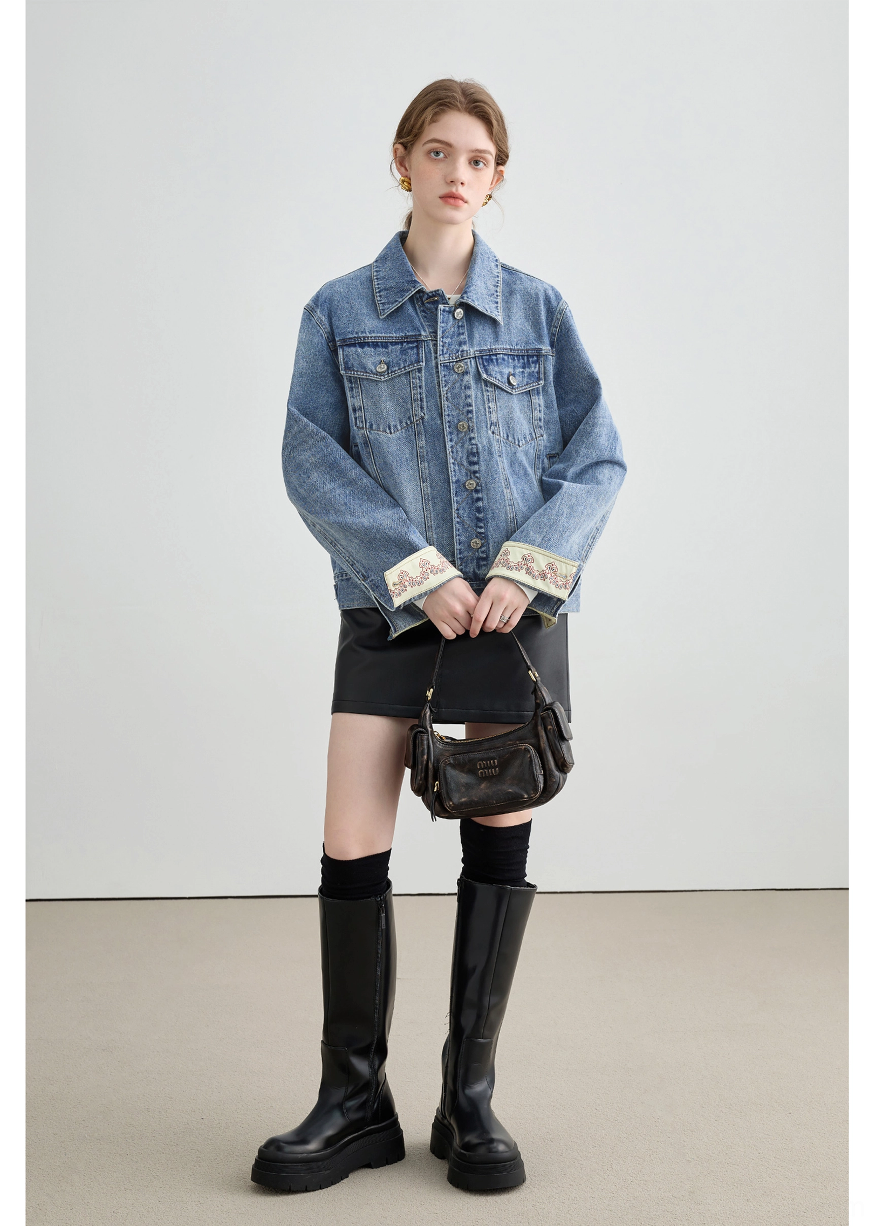Side front view of a female model in a relaxed light wash denim jacket with embroidered turn-back cuffs and black mini skirt
