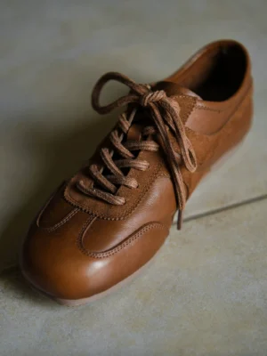 Warm brown retro leather sneakers with suede lace panel, rounded toe, tonal laces, and low-top streetwear silhouette photographed on a beige tiled floor