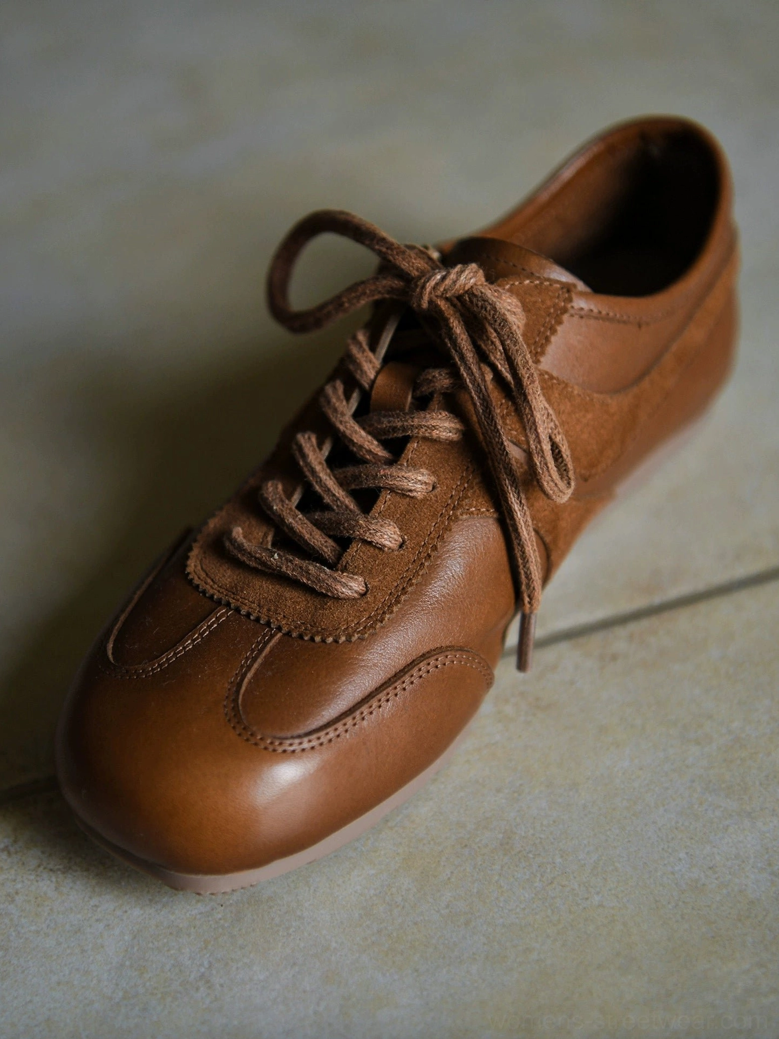 Warm brown retro leather sneakers with suede lace panel, rounded toe, tonal laces, and low-top streetwear silhouette photographed on a beige tiled floor