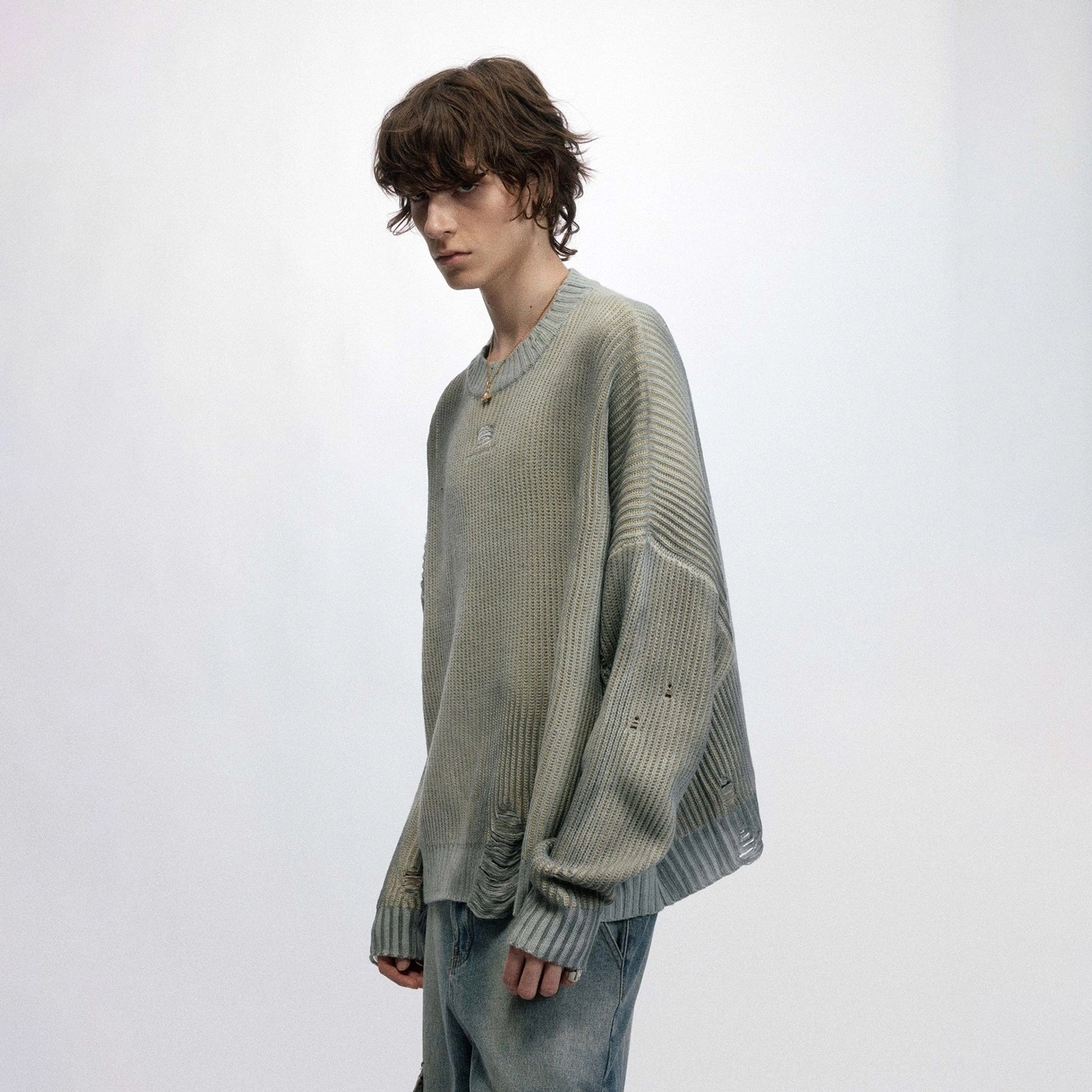 Three-quarter view of a male model wearing a sage gray distressed ribbed sweater with drop shoulders and denim trousers