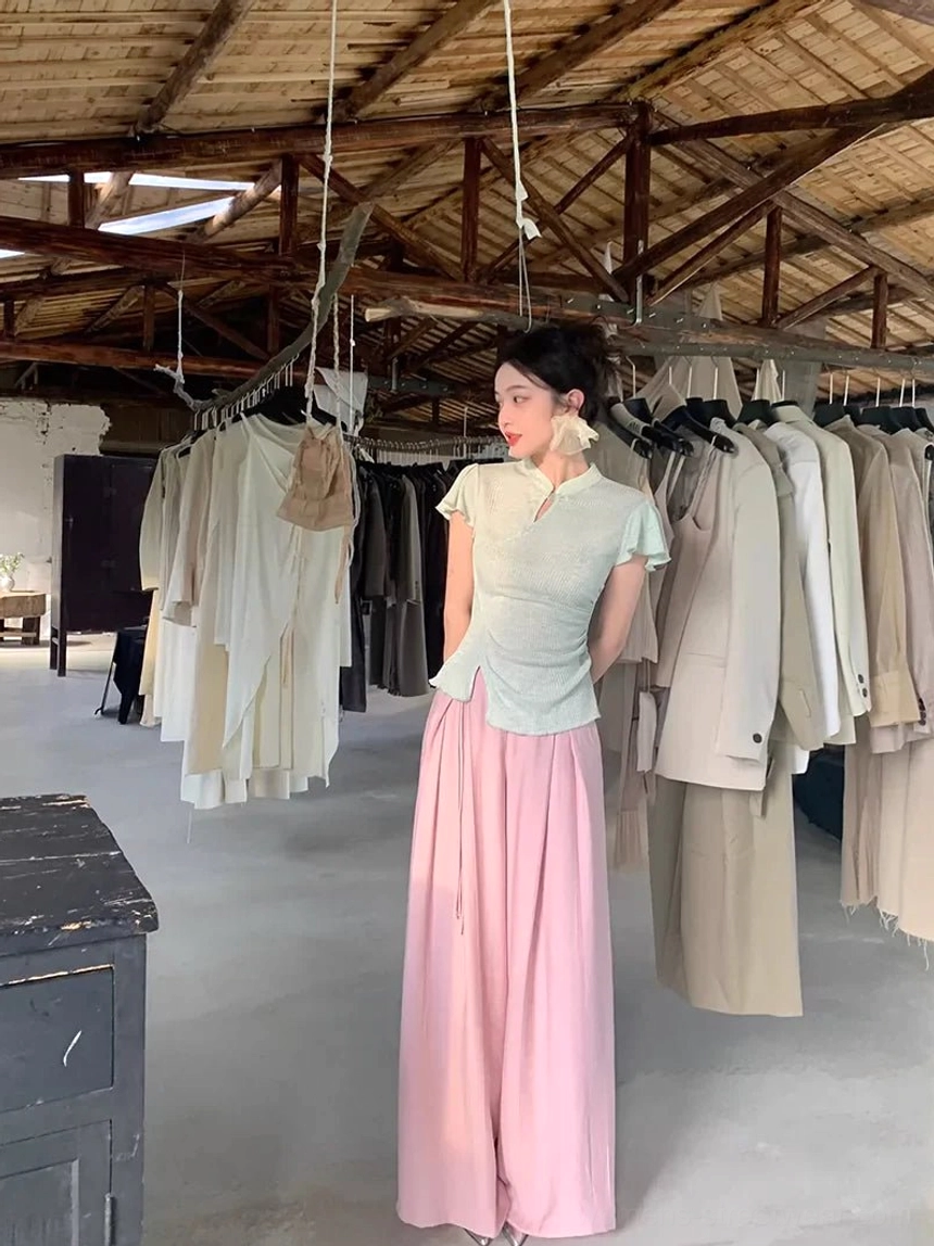 Side three-quarter view of a model wearing a fitted sage green asymmetrical cheongsam top with a soft pink high-waisted flowing skirt