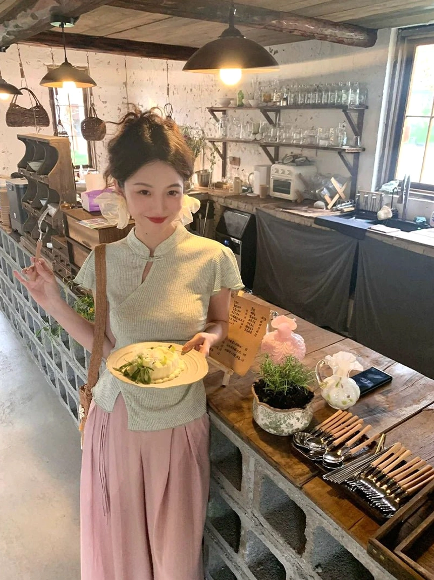 Model wearing a sage green asymmetrical ribbed cheongsam top with ruffled sleeves and a flowing pink maxi skirt in a retro cafe setting
