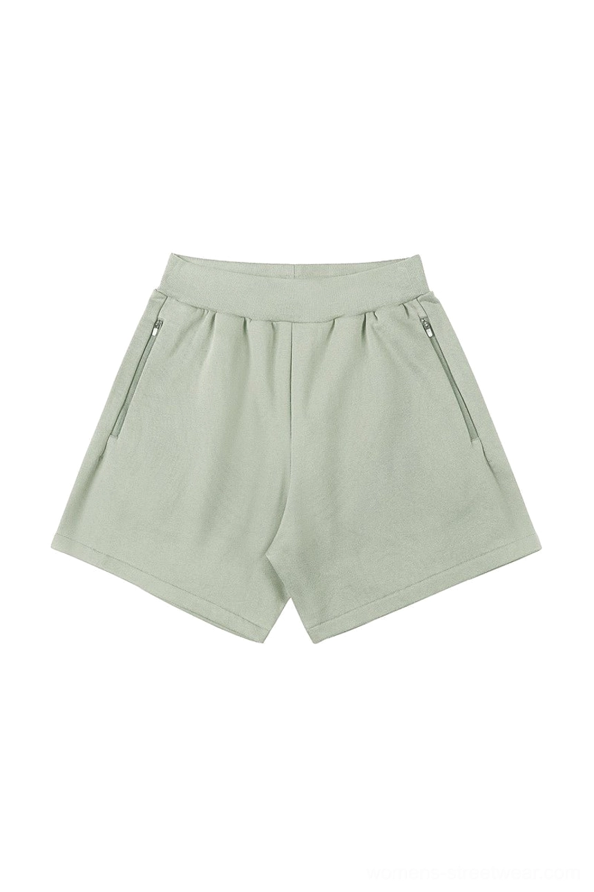 Muted sage green cotton blend streetwear shorts with elastic ribbed waistband and side zipper pockets shown on a white background