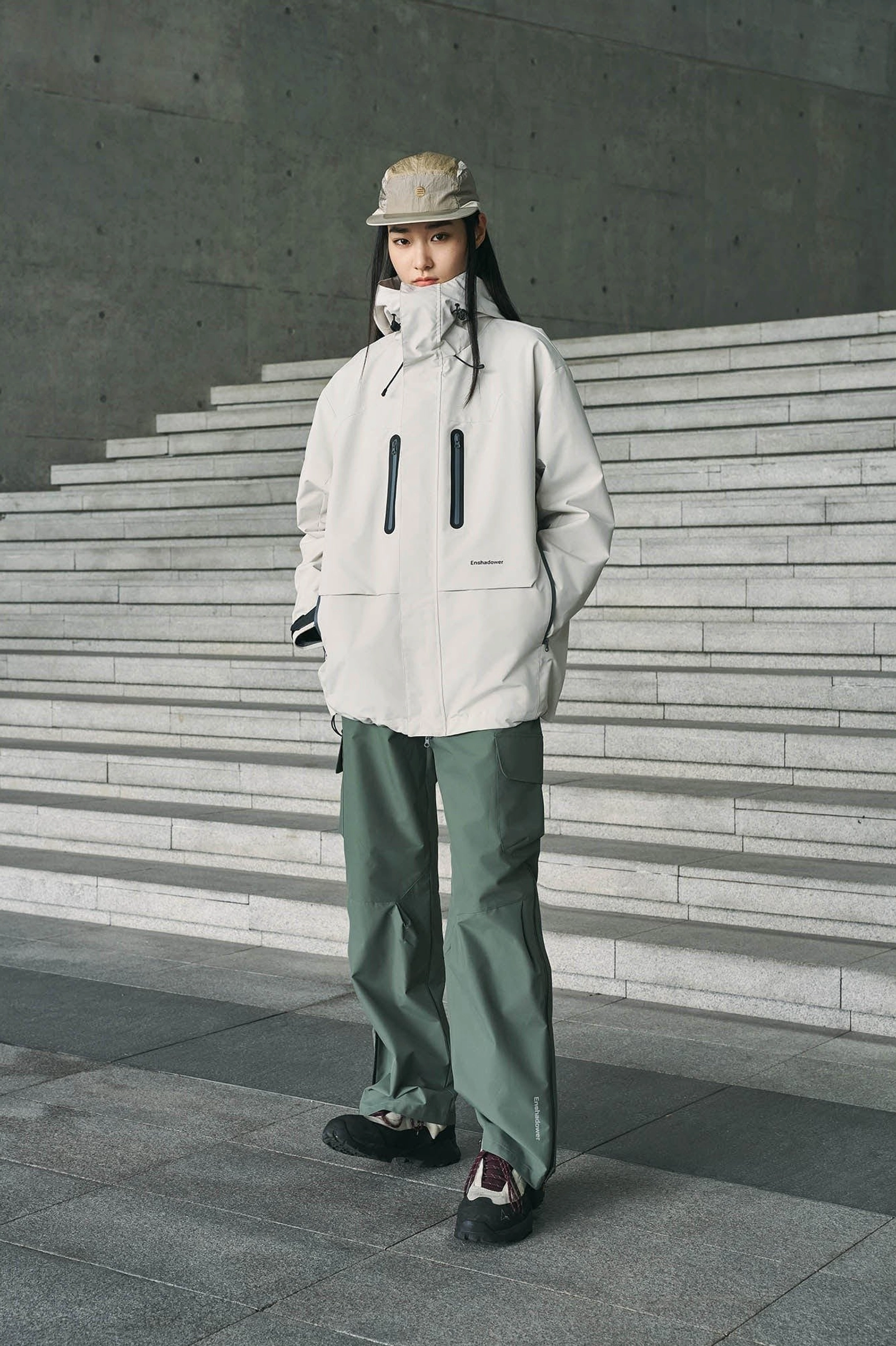 Sage green techwear cargo pants paired with a neutral grey outdoor jacket and bucket hat for a modern urban gorpcore look