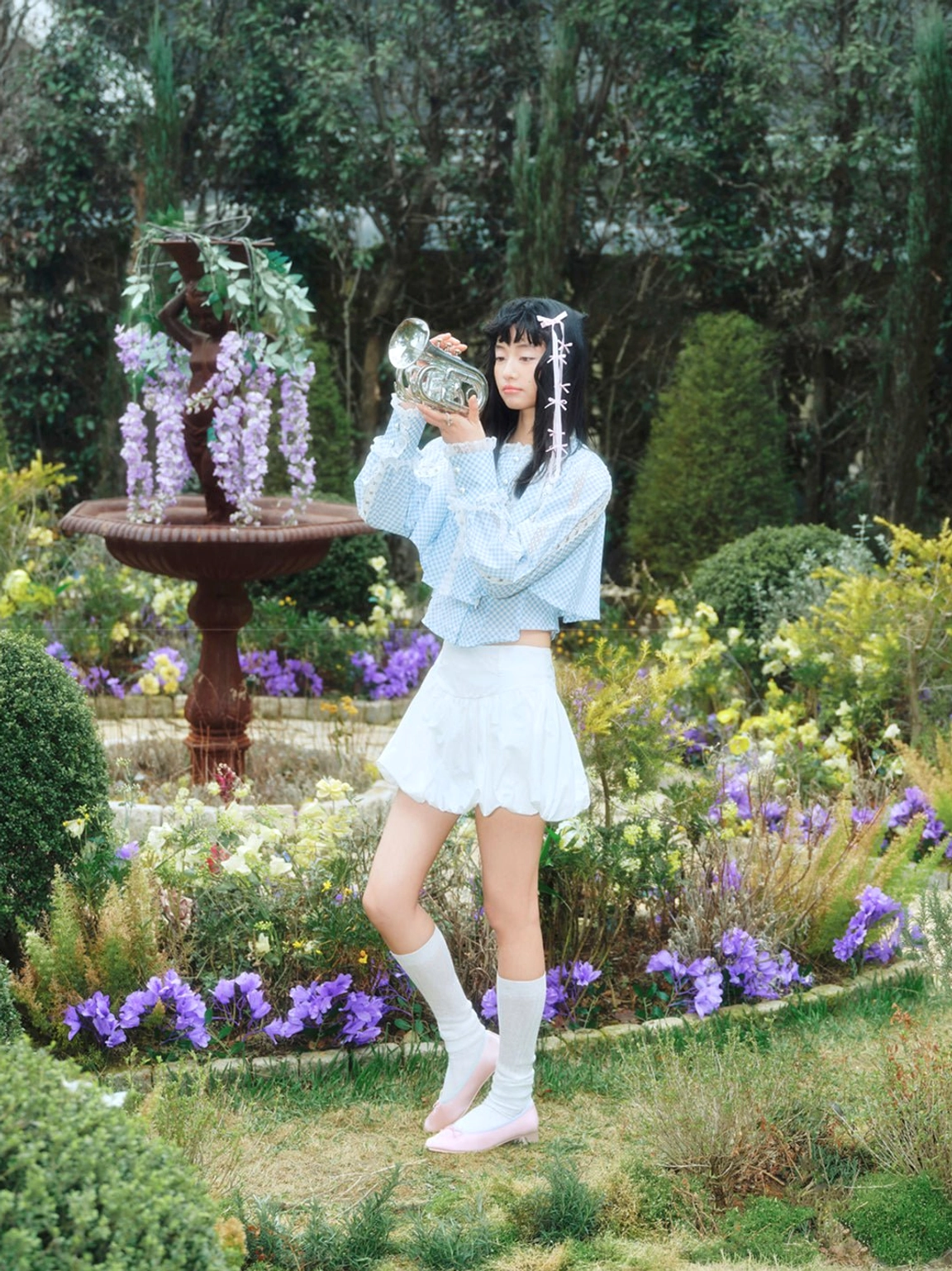Three-quarter fashion view of pink satin bow ballet pumps styled with a blue cropped top and garden backdrop