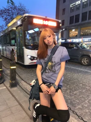 Female model wearing a dusty blue Scream Kids washed gradient graphic tee with a distressed front print on an urban night street beside a public bus