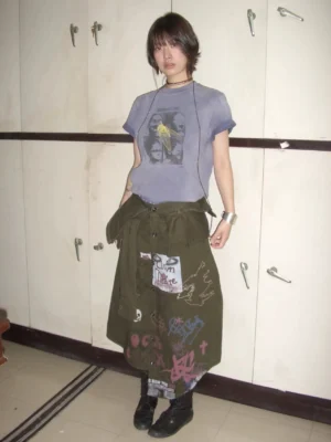 Full body female model wearing a washed purple blue Scream Kids graphic T-shirt styled with a printed cargo skirt in a locker room
